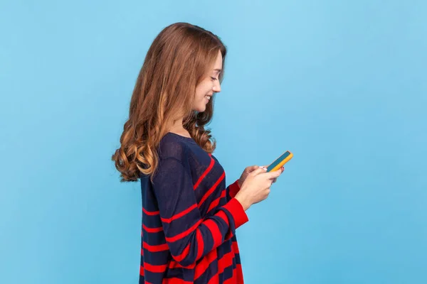 笑顔の楽観的な女性が手にスマートフォンで立って ボーイフレンドからロマンチックなメッセージに答える 縞模様のカジュアルなセーターを着て 屋内スタジオは青の背景に隔離されたショット — ストック写真