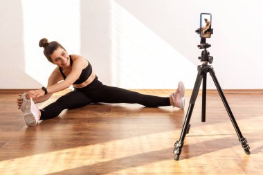 Kız koç bacak esnetiyor, internet üzerinden yayın yapıyor, video kaydı için telefon kullanıyor, siyah spor bluzu ve tayt giyiyor. Pencereden gelen güneş ışığıyla aydınlatılan tam uzunluktaki stüdyo görüntüleri..