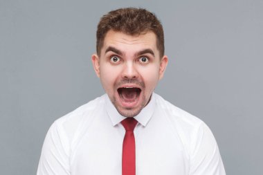 Beyaz gömlekli, kravatlı, şok olmuş, ağzı açık kameraya bakan genç ve yakışıklı bir adamın portresi. Gri arkaplanda kapalı alan.
