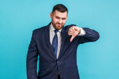 Resmi stil takım elbise giyen, başparmağını aşağı gösteren ve kameraya bakan tatminsiz sakallı bir adamın portresi. Uygulama testi geribildirimi. Mavi arka planda kapalı stüdyo çekimi.