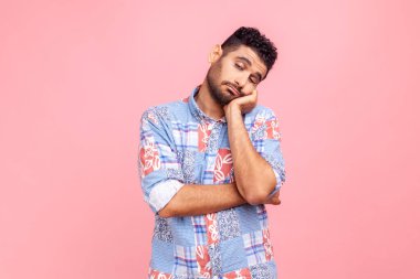 Mavi tişörtlü, sakallı, kafası el tembelliği, ilgisizlik ve tembellik üzerine eğilmiş canı sıkkın bir adamın portresi uykulu görünüyor. Pembe arka planda kapalı stüdyo çekimleri.