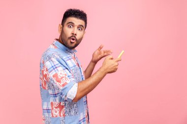 Şok olmuş bir ifadeyle cep telefonunu işaret eden genç bir adamın yan görüntüsü, büyük gözleri ve ağzı açık kameraya bakan, pembe arka planda izole edilmiş kapalı stüdyo görüntüsü..