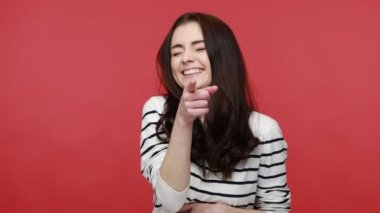 Komik kadın portresi yüksek sesle gülüyor, kamerayı işaret ediyor, gülünç bir aptalı gösteriyor, uzun kollu bir gömlek giyiyor. Kırmızı arkaplanda kapalı stüdyo çekimleri.