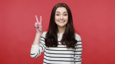 Ziyaretçi işareti gösteren bir kadın, zaferinden memnun, kameraya dişlek bir gülümsemeyle bakıyor, başarı, günlük tarz uzun kollu gömlek giyiyor. Kırmızı arkaplanda kapalı stüdyo çekimleri.