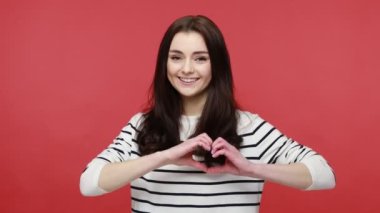 Elleriyle kalp şekli yapan, kameraya dostça gülümseyen, sevgi ve özen gösteren, gündelik tarzda uzun kollu gömlek giyen bir kadın. Kırmızı arkaplanda kapalı stüdyo çekimleri.