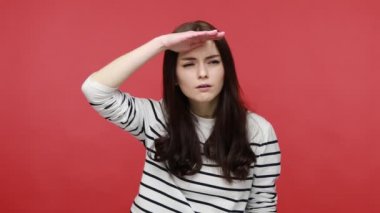 Kadın avuçlarını başının üstünde tutuyor, dikkatle uzaklardan bakıyor, uzun kollu sıradan bir gömlek giymiş, uzun mesafeli beklentilerle bakıyor. Kırmızı arkaplanda kapalı stüdyo çekimleri.