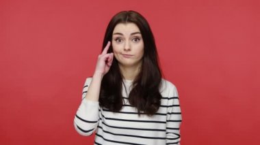 Portrait of woman making stupid sign with finger near head, gesturing bad mind, dumb insane idea, wearing casual style long sleeve shirt. Indoor studio shot isolated on red background.