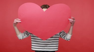 Romantik esmer kadının portresi, büyük bir kalp aşk sembolü taşıyor, hava öpücüğü veriyor, flört ediyor, günlük tarz uzun kollu gömlek giyiyor. Kırmızı arkaplanda kapalı stüdyo çekimleri.