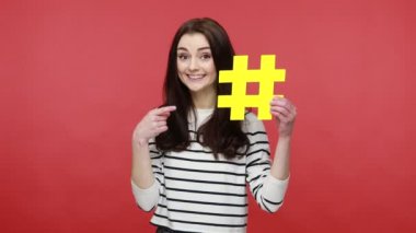 Kadın sarı hashtag sembolünü işaret ediyor, foruma katılmayı öneriyor, popüler blog fikri, günlük tarz uzun kollu gömlek giyiyor. Kırmızı arkaplanda kapalı stüdyo çekimleri.