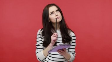 Düşünceli bir kadının portresi not defterine kalemle düşünceler yazmak, liste yapmak, günlük tarz uzun kollu gömlek giymek. Kırmızı arkaplanda kapalı stüdyo çekimleri.