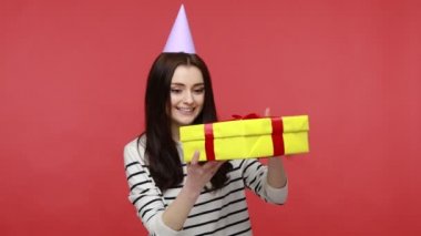 Parti konisi içinde doğum gününü kutlayan bir kadın portresi hediye kutusunu açıyor, hoş bir sürpriz alıyor, günlük tarz uzun kollu gömlek giyiyor. Kırmızı arkaplanda kapalı stüdyo çekimleri.