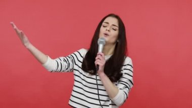 Çekici bir kadının portresi, elinde mikrofonla dans etmek, neşeli şarkılar söylemek, uzun kollu bir gömlek giymek. Kırmızı arkaplanda kapalı stüdyo çekimleri.