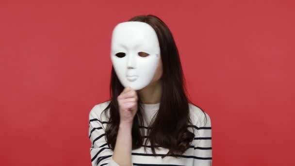 若いブルネットの女性の肖像は カジュアルスタイルの長袖シャツを着て 異なる感情 偽善を示すマスクを脱ぎます 屋内撮影所は赤の背景に隔離された — ストック動画