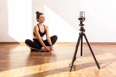 Kadın koç idmandan sonra dinlenme videosu çekiyor, Lotus 'un üzerinde gözleri kapalı oturuyor, siyah spor bluzu ve tayt giyiyor. Pencereden gelen güneş ışığıyla aydınlatılan tam uzunluktaki stüdyo görüntüleri..
