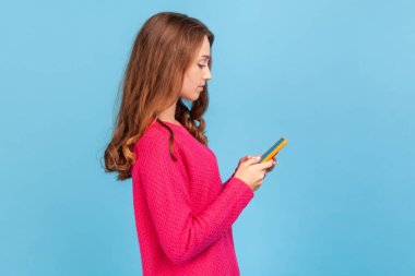 Kenara çekilen ciddi bir kadının yan görüntüsü, cep telefonuyla yazı yazması, blog okuması, sosyal ağda sohbet etmesi, internet iletişimi için mobil uygulama kullanması. Mavi arka planda kapalı stüdyo çekimi.