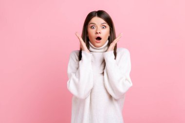 Şok olmuş, koyu renk saçlı, kollarını kaldırmış, kameraya açık ağızlı beyaz süveter giymiş bir kadın. Pembe arka planda kapalı stüdyo çekimleri.