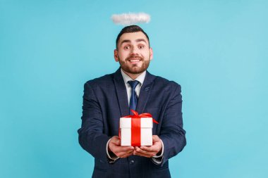 Sakallı, nazik gülümsemeli resmi stil takım elbiseli ve kafasının üzerinde haleler olan bir adam. Hediye kutusu, hayır kurumu, din, kameraya bakmak. Mavi arka planda kapalı stüdyo çekimi.