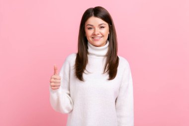 Gülümseyen esmer kadın kameraya baş parmağını gösteriyor, onaylama ve olumlu duygularını ifade ediyor, beyaz günlük süveter giyiyor. Pembe arka planda kapalı stüdyo çekimleri.