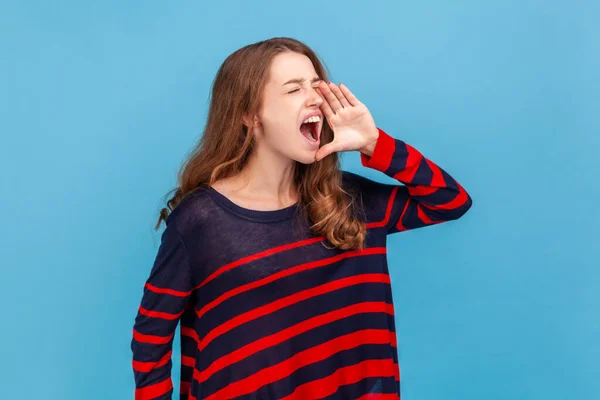 怒っている女性の縞模様のカジュアルなスタイルのセーターを大声で叫んで 口の近くに手を取り 重要なニュースを発表し 注目を集めて身に着けている 青い背景に隔離された屋内スタジオショット — ストック写真