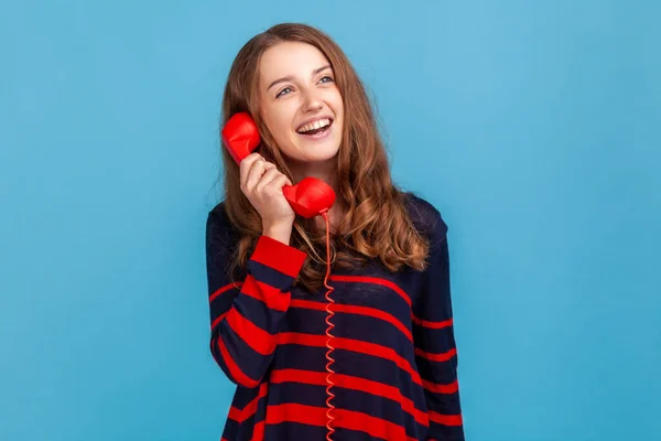 年轻的成年女性穿着条纹休闲装 通过红色复古固定电话交谈 进行愉快的交谈 面带微笑地向别处看去 室内拍摄被蓝色背景隔离 — 图库照片