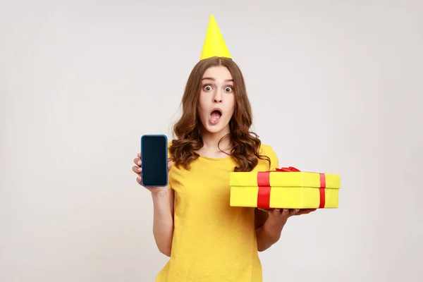 不可信的手机用户生日奖金 惊慌失措的少女在生日圆锥上拿着礼品盒和带空白显示屏的手机的画像 在灰色背景下被隔离的室内拍摄 — 图库照片