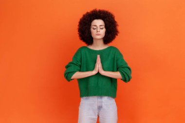 Sakin, Afro saç stili, yeşil rahat süveter giyen, yoga yapan, nefes egzersizi yapan, rahat, genç bir kadın stresi azaltıyor. Turuncu arka planda kapalı stüdyo çekimleri.