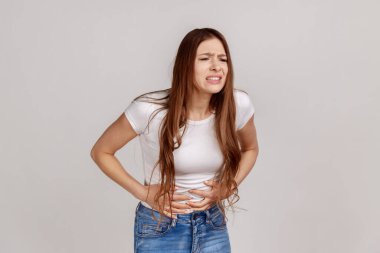 Karnını tutan, akut karın ağrısı, gastrit ya da kabızlık yüzünden yüzünü buruşturan, beyaz tişört giyen sağlıksız bir kadının portresi. Gri arka planda izole edilmiş kapalı stüdyo görüntüleri.