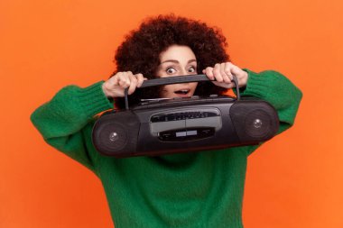 Afro saçlı, yeşil kazak giyen, elinde kayıt cihazıyla kameraya heyecanlı bir ifadeyle bakan mutlu, komik bir kadın. Turuncu arka planda kapalı stüdyo çekimleri.