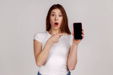 Cep telefonunu işaret eden ve şaşırmış bir ifadeyle beyaz tişört giyen, gezici tarifelerden şok olmuş genç bir kadının portresi. Gri arka planda izole edilmiş kapalı stüdyo görüntüleri