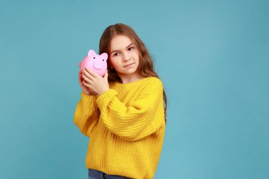 Çekici, meraklı küçük bir kızın portresi, elinde domuz para kutusu, tasarruf, finansal ya da bankacılık, sarı gündelik süveter giyiyor. Mavi arka planda kapalı stüdyo çekimi.