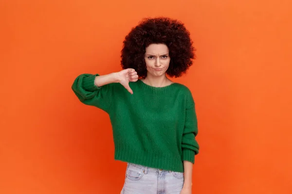 親指ダウン 悪いサービスやコンテンツを示す緑のカジュアルなスタイルのセーターを着てアフロの髪型と悲しい落ち込んで女性は お勧めしません オレンジの背景に隔離された屋内スタジオショット — ストック写真