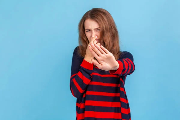 Woman wearing striped sweater grabbing nose, grimacing in disgust and showing stop gesture, expressing repulsion to stink, fart gases, intolerable odor. Indoor studio shot isolated on blue background.