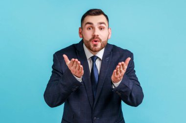 Resmi tarz kıyafet giymiş sakallı genç bir adam avuç içlerine hava öpücüğü gönderiyor, kameraya flört ediyor, romantik bir aşk yaşıyor. Mavi arka planda kapalı stüdyo çekimi.