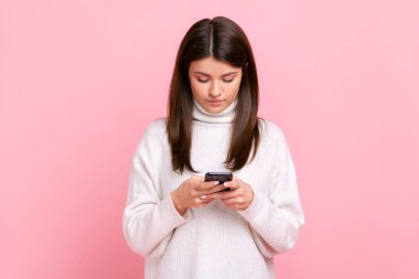 Akıllı telefon kullanan, sosyal ağları kontrol eden güzel bir kadının portresi, beyaz günlük süveter giyen, konsantre bir ifadeye sahiptir. Pembe arka planda kapalı stüdyo çekimleri.