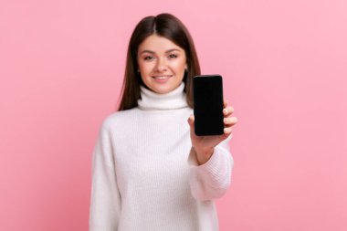 Gülümseyen esmer kadın reklam veya tanıtım metni için boş ekranlı akıllı telefon gösteriyor, beyaz günlük tarzda süveter giyiyor. Pembe arka planda kapalı stüdyo çekimleri.