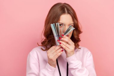 Kapüşonlu, meraklı, kıvırcık saçlı genç kız portresi bir sürü dolar banknotunu dikizliyor, pasif gelir. Pembe arka planda kapalı stüdyo çekimleri