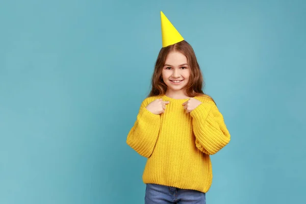 パーティーコーンの女の子はカメラを見て笑顔で 自分自身を指して 誕生日を祝い 黄色のカジュアルなスタイルのセーターを着ています 青い背景に隔離された屋内スタジオショット — ストック写真