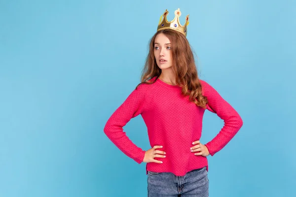 Arrogant woman wearing pink pullover, in crown on head looking away with confident expression, self-motivation and dreams to be best. Indoor studio shot isolated on blue background.