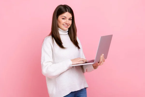 幸せな顔をしたカメラを見て成功したオフィスワーカーの女の子は キーボードを入力し 白いカジュアルなスタイルのセーターを着てノートパソコンを保持 ピンクの背景に隔離された屋内スタジオショット — ストック写真