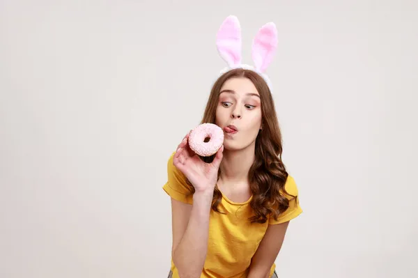 Kahverengi Saçlı Donut Yalamak Için Dilini Gösteren Pembe Tavşan Kulaklı — Stok fotoğraf