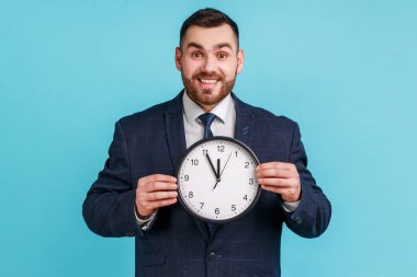 Büyük bir saat tutan koyu renk takım elbiseli, pozitif neşeli iş adamının portresi. Ve kameraya dişlek gülümsemeli, iyimser bir ifadeyle bakıyor. Mavi arka planda kapalı stüdyo çekimi.