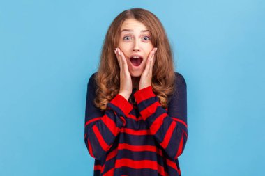 Buna inanamam. Şok olmuş bir ifadeyle kameraya bakan süveter giyen, gülümseyen ve el sallayan bir kadın, harika haberler, zafer. Mavi arka planda kapalı stüdyo çekimi.