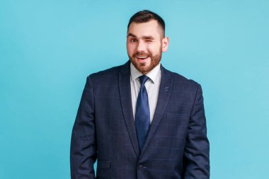 Resmî stil takım elbiseli, sakallı iyimser genç adam kameraya bakıyor ve göz kırpıyor, olumlu duygularını ifade ediyor. Mavi arka planda kapalı stüdyo çekimi.