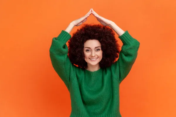 アフロの髪型家の屋根のジェスチャーや笑顔 生命保険 保護の概念を作る緑のカジュアルなスタイルのセーターを着て女性 オレンジの背景に隔離された屋内スタジオショット — ストック写真