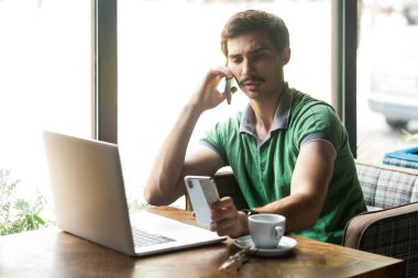 Yeşil tişört giyen, dizüstü bilgisayarda çalışan, cep telefonuyla konuşan ve başka bir cep telefonuna bakan yakışıklı işadamının portresi. Büyük pencerenin yanında, kafenin arka planında..