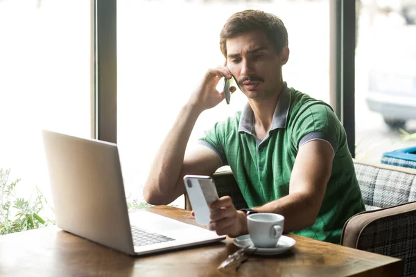緑のTシャツを着た忙しいハンサムなビジネスマンの肖像画 ラップトップで作業し 携帯電話で話し 別の携帯電話を見ています 大きな窓の近くの屋内撮影 カフェの背景 — ストック写真
