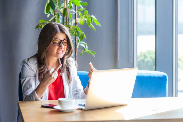 Грубая женщина в очках, показывающая средний палец на экране ноутбука, агрессивно общающаяся по видеосвязи, выражающая неуважение и ненависть. Крытый снимок, кафе или офисный фон.