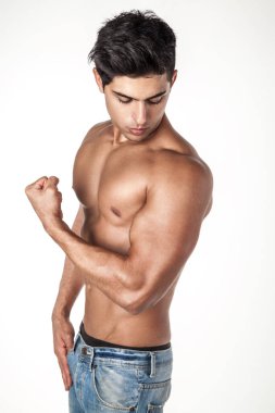 Koyu renk saçlı, kot pantolon giyen, güçlü kolunu kameraya gösteren ve pazularına bakan genç bir adamın yan görünüm portresi. Beyaz arka planda izole edilmiş kapalı stüdyo görüntüleri.