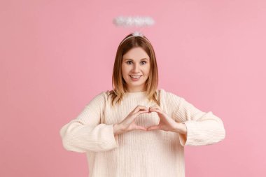 Başında nimbüsle kameraya bakıp kalp şeklini elleriyle gösteren beyaz süveter giyen sevimli sarışın meleğin portresi. Pembe arka planda kapalı stüdyo çekimleri.
