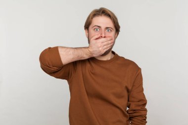 Sakallı, kazaklı korkmuş bir adamın portresi, ağızdan ağıza, korkudan şok olmuş gözlerle bakıyor, gizli bir tabu tutuyor. Gri arka planda izole edilmiş kapalı stüdyo görüntüleri.